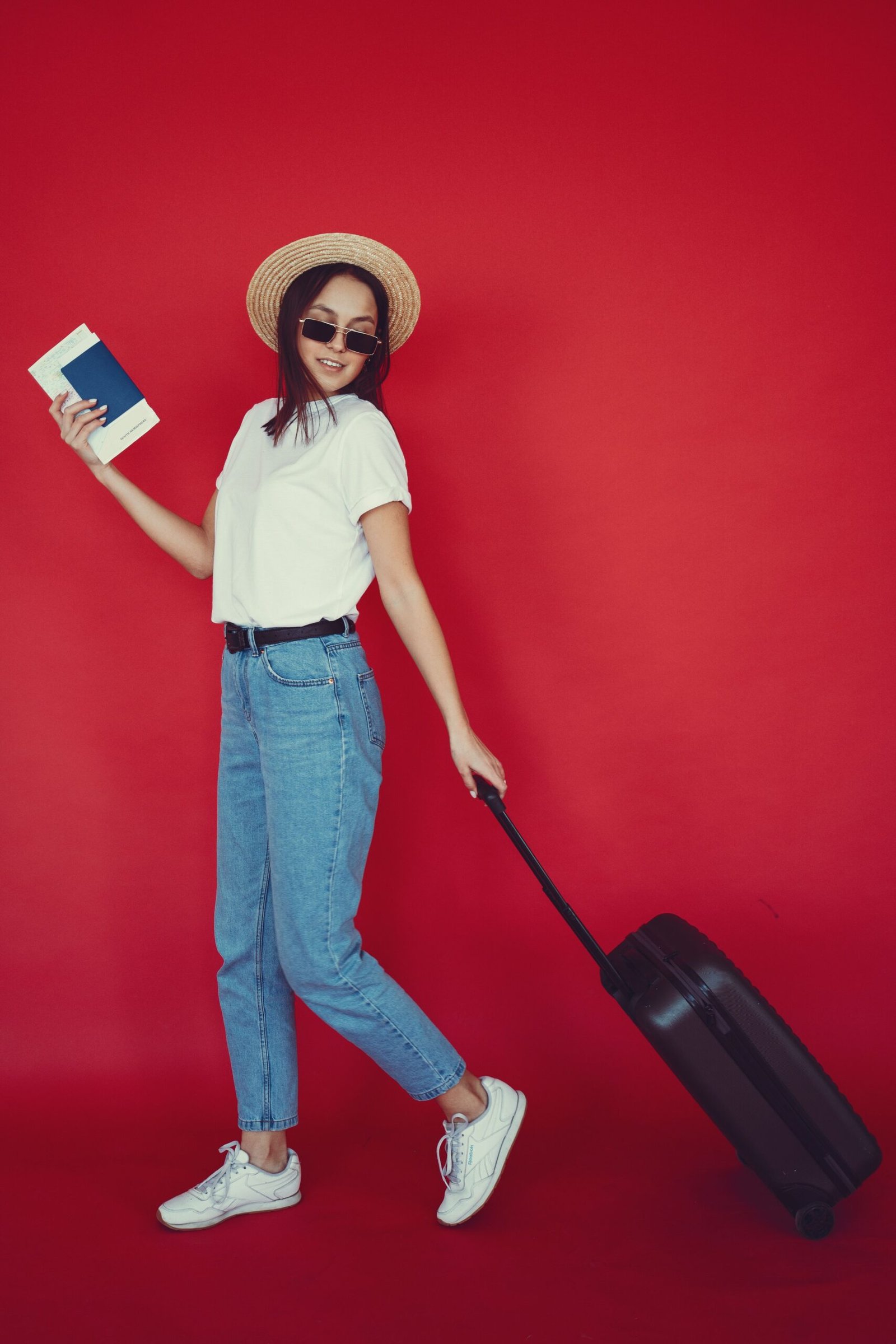 Full body young woman with long hair in jeans carrying luggage and looking at camera while holding passport and tickets on red background